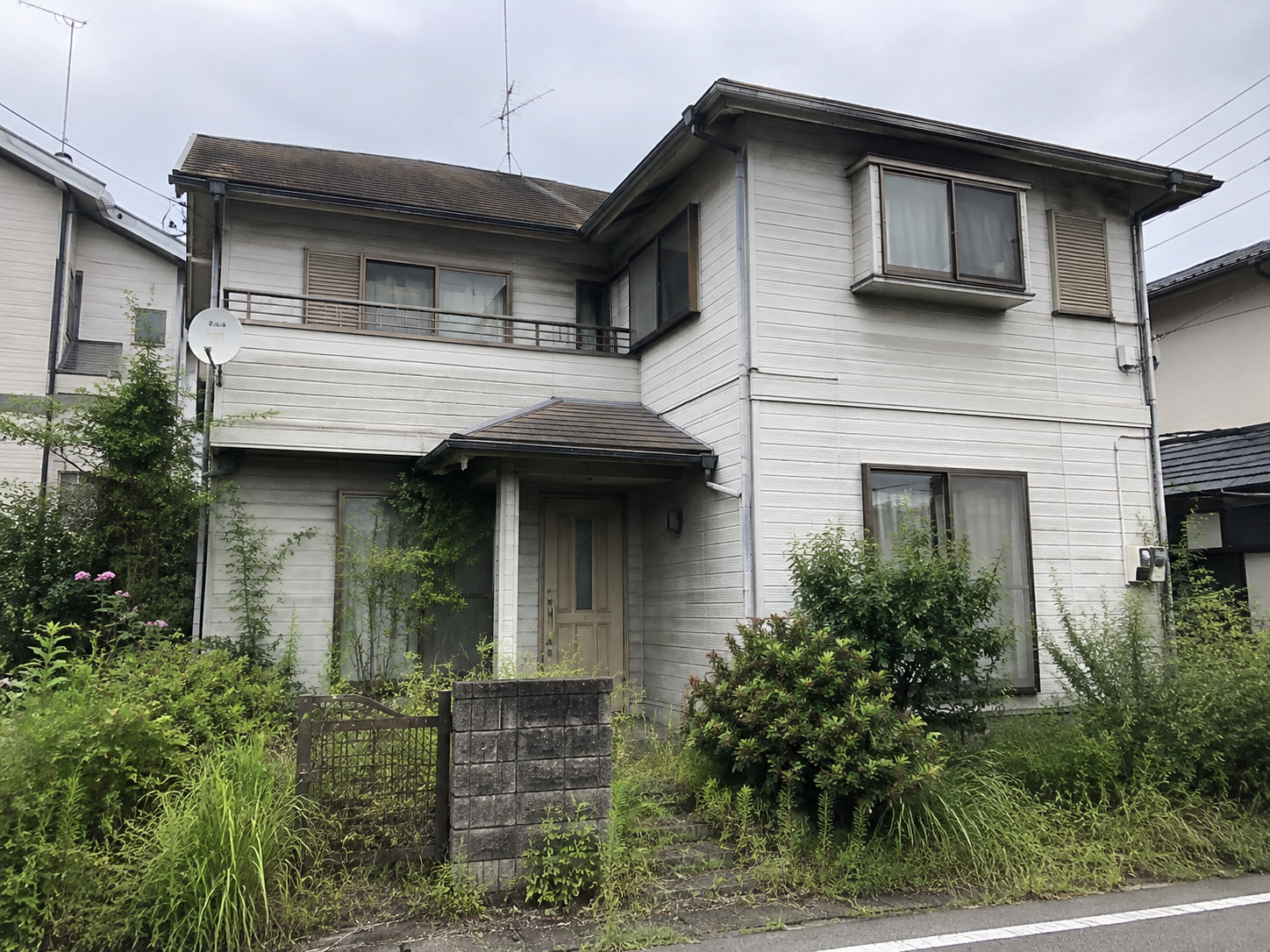 経年劣化した外壁・植栽が伸びた庭を持つ、築25〜30年程度の一戸建て住宅の外観写真